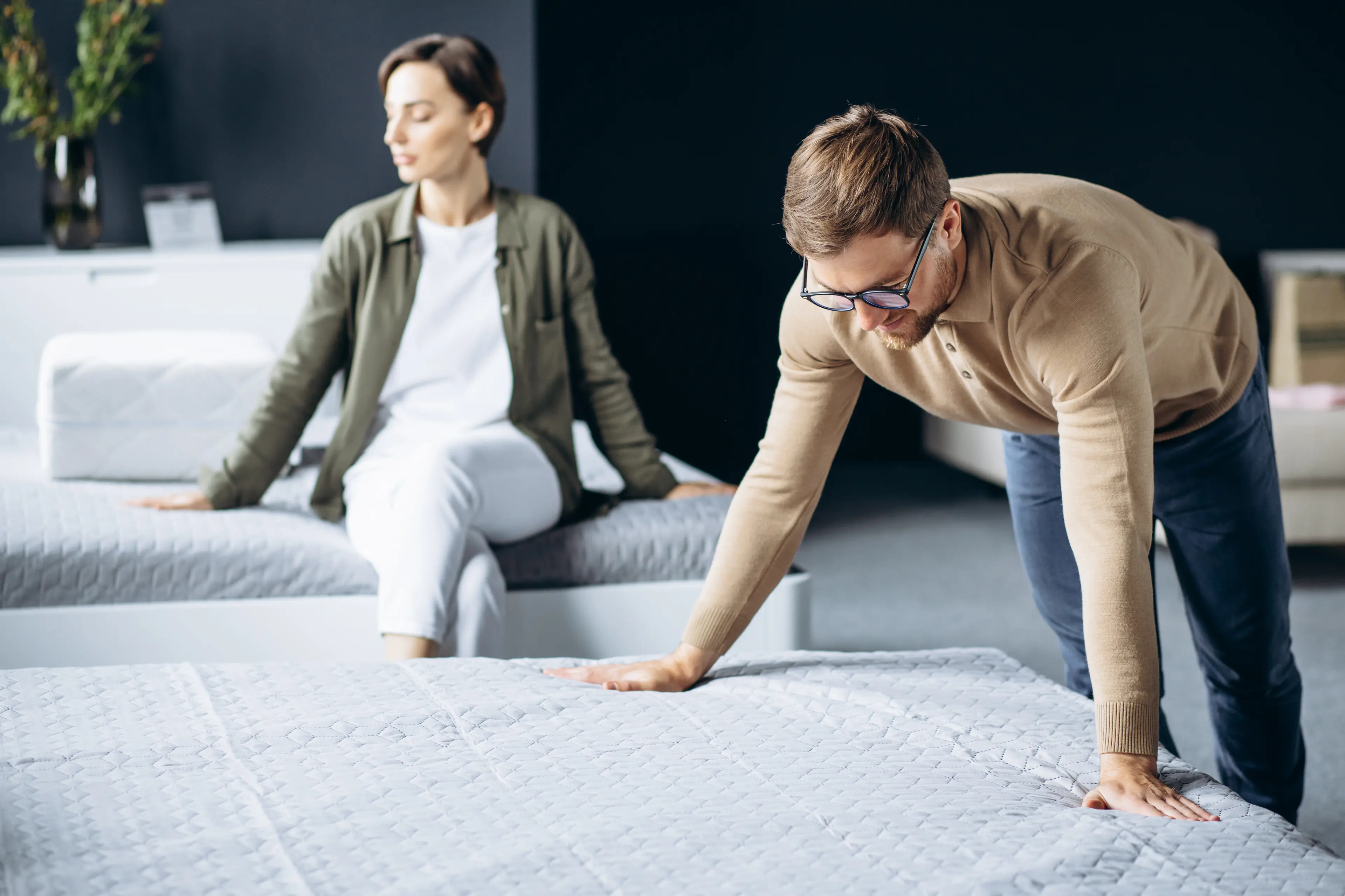 A man and woman test mattresses in a bright store. He leans forward, both hands on the mattress to test the firmness while the woman is sitting on another mattress to test the comfort. They are not engaged, perhaps zoned in their own mattress experiences.